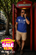 Man wearing a royal blue Rick's Café Américain graphic t-shirt with white palm tree print, standing in a red London telephone box